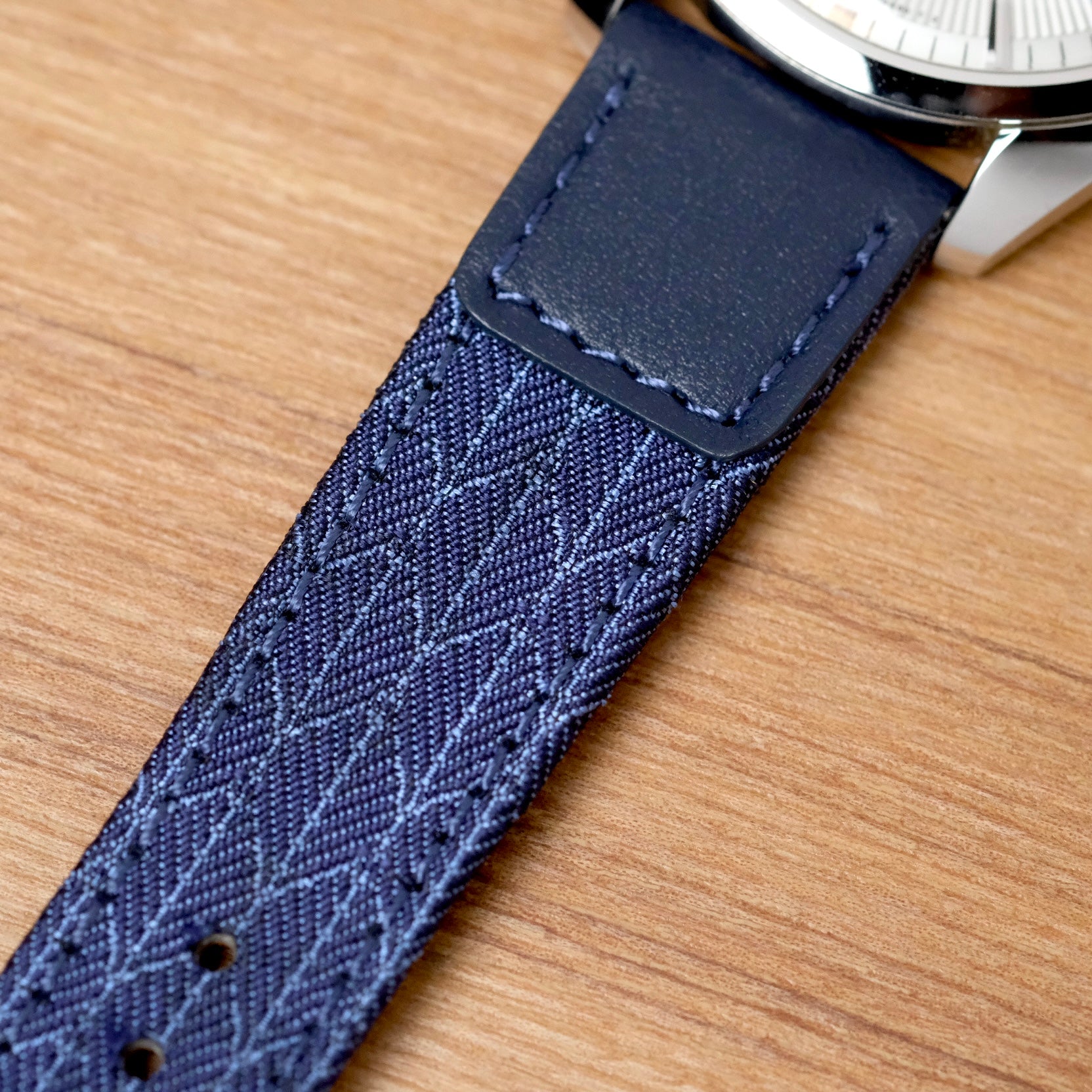 Close-up of the RSM Watch Strap’s Art Deco Silk Watch Strap in Blue, featuring a geometric textured fabric, attached to a metal watch case and placed on a wooden surface.