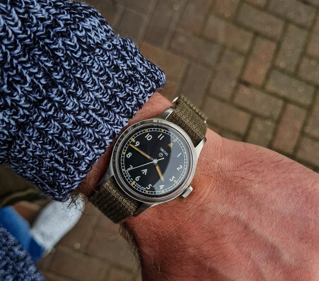 The VTG Mil in Green by RSM Watch Strap, with a black-dial watch and green fabric strap, pairs seamlessly with a blue sweater.