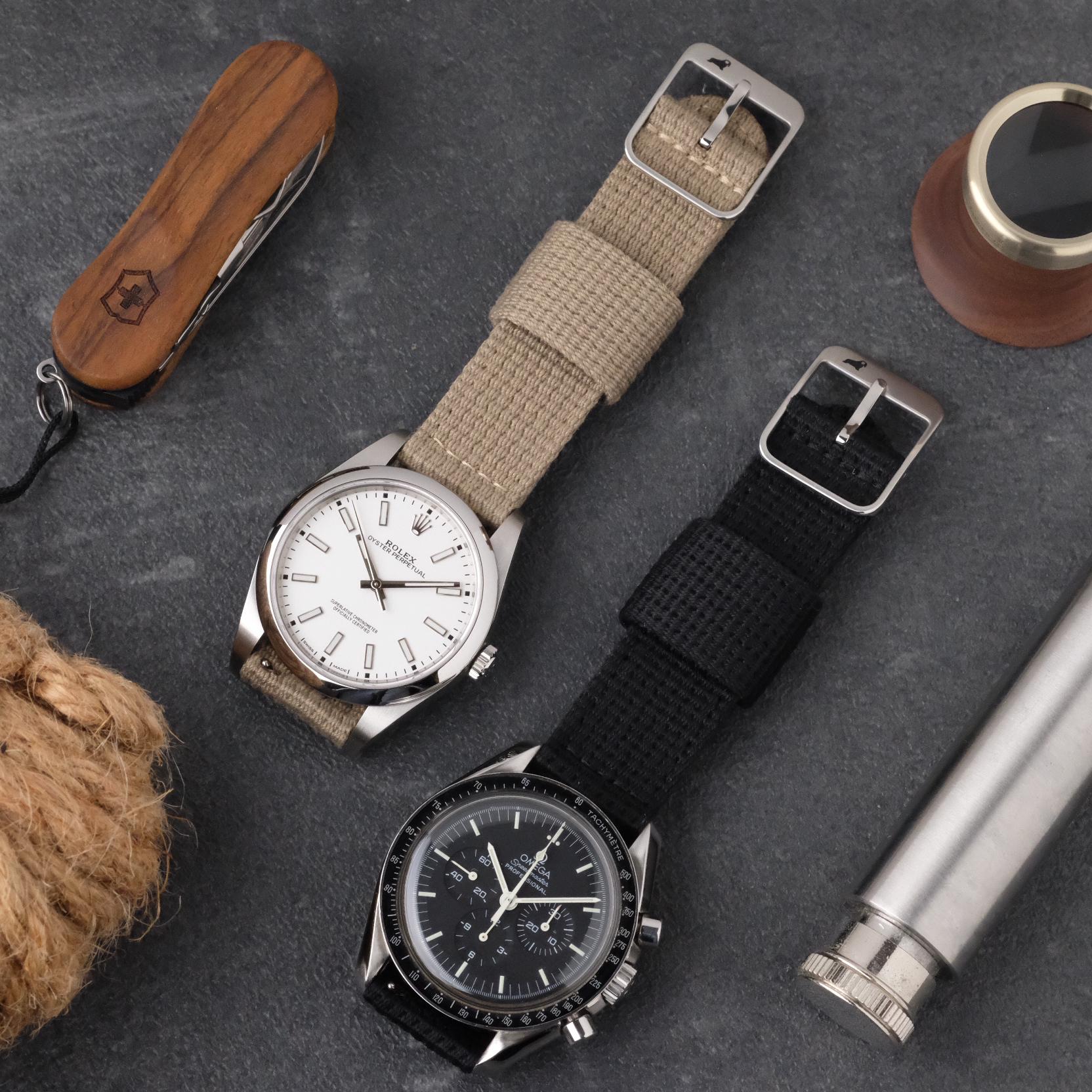 Two watches fitted with RSM Watch Strap’s Vintage Military 2-Piece Watch Strap in Steel—one beige with a white dial, one black with a black dial—rest on a gray surface alongside a wooden-handled knife, rope, metal cylinder, and small magnifier.
