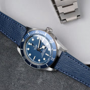 A Tudor dive watch with a blue bezel, round hour markers, and white hands sits on a grey surface paired with an RSM Watch Strap Italian Epsom Leather Watch Strap in Navy Blue, next to a stainless steel bracelet.