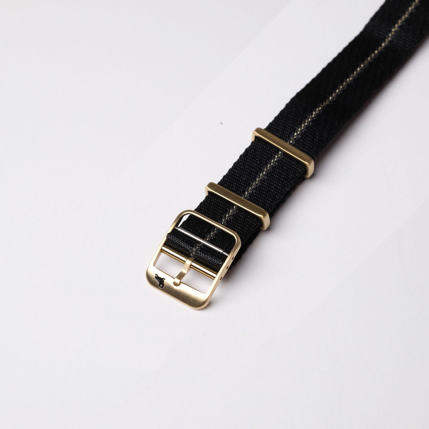 The RSM Watch Strap Engraved Stripe in Gilt Stripe features a black fabric belt with a brass buckle, two loops, and a Jacquard weave, laid on white.
