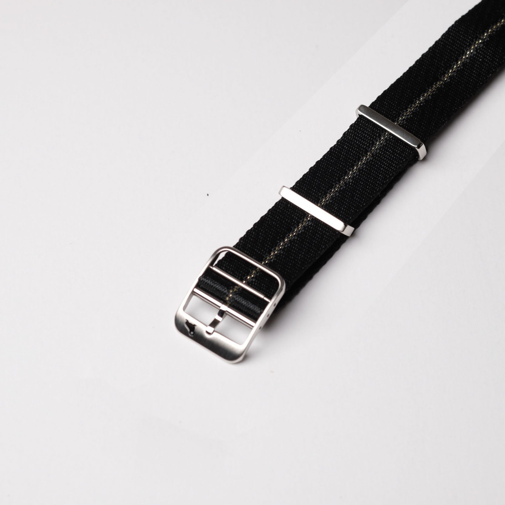 The RSM Watch Strap Engraved Stripe in Gilt Stripe features a black nylon strap, silver buckle, loops, and a subtle thin stripe on a gray background.