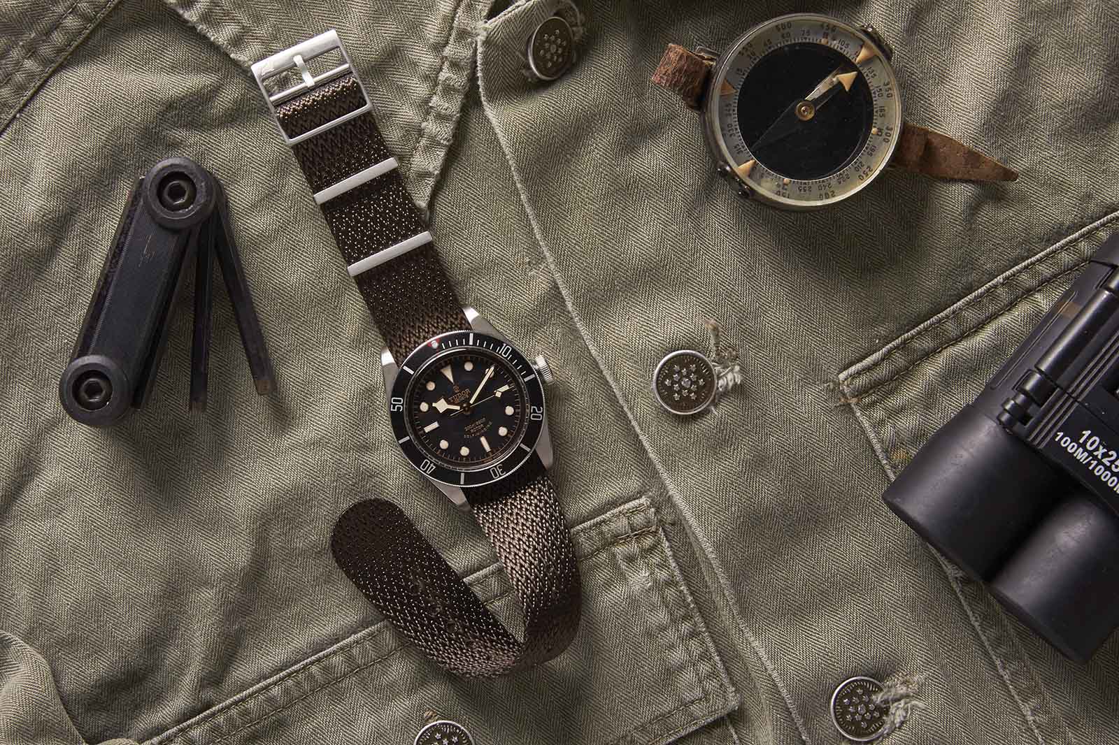 RSM Herringbone Twill Tobacco Brown strap rests on a green military jacket with a compass, multi-tool, and binoculars, evoking an adventurous vibe.