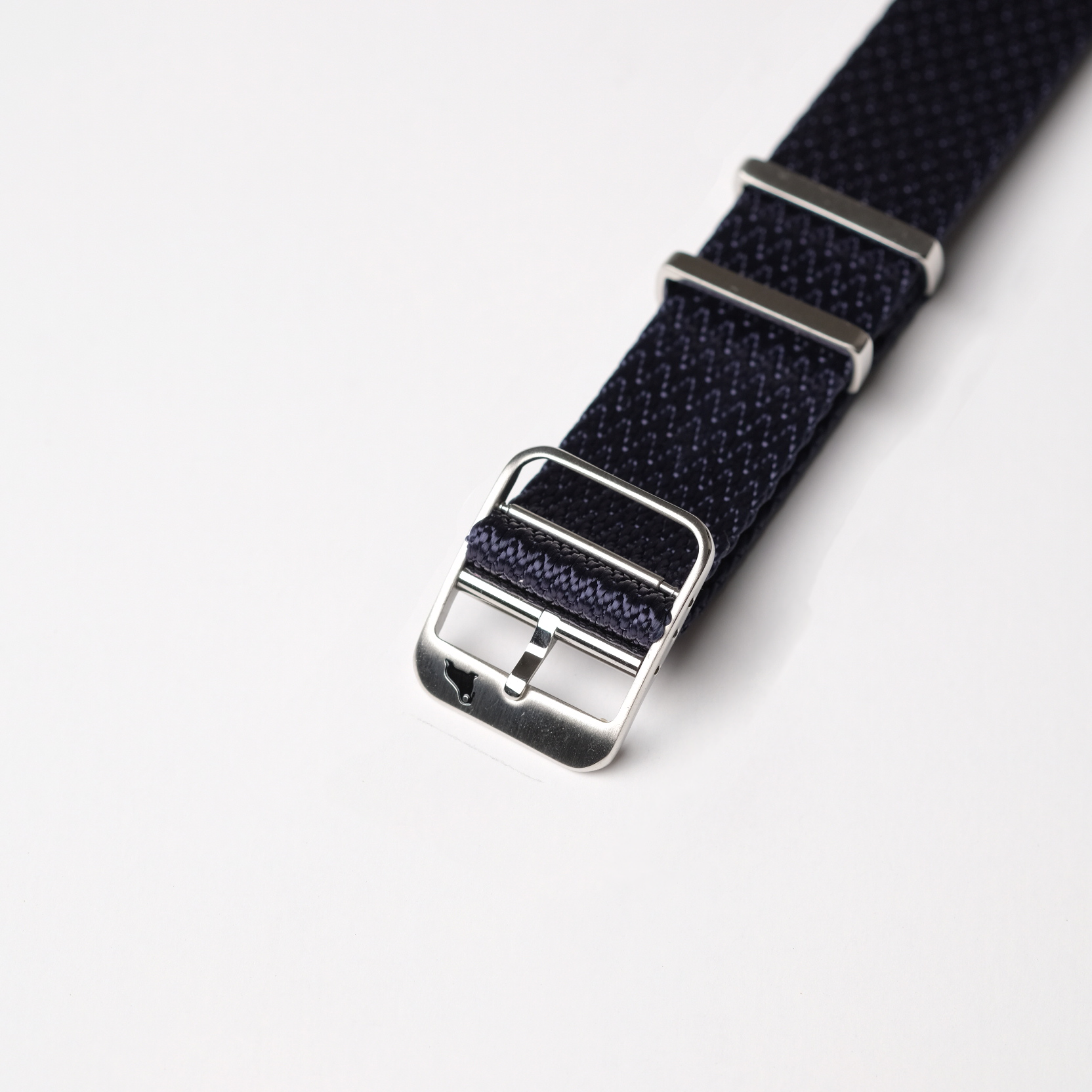Close-up of RSM Watch Straps Herringbone Twill in Icarus Blue, featuring silver metal buckles and loops on a white surface, highlighting the intricate texture.