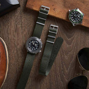 A flat lay features two watches with RSM Watch Straps Herringbone Twill in Olive Drab, wooden board, and aviator sunglasses.
