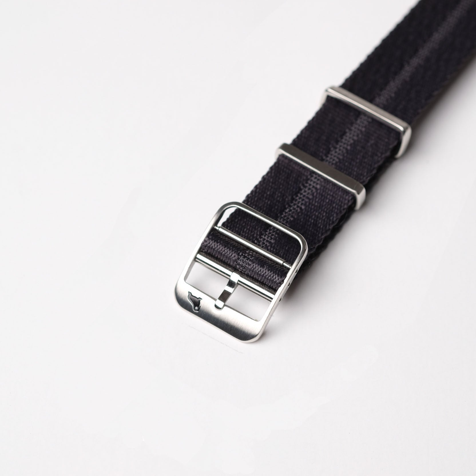 Close-up of RSM's Engraved Stripe in Stealth Gray, a black fabric strap with silver buckle, metal loops, and discreet logo on white.