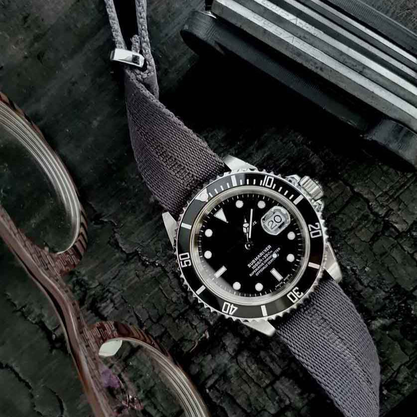 The Engraved Stripe in Stealth Gray by RSM Watch Strap features a silver and black divers watch with adjustable fabric band, resting on wood.