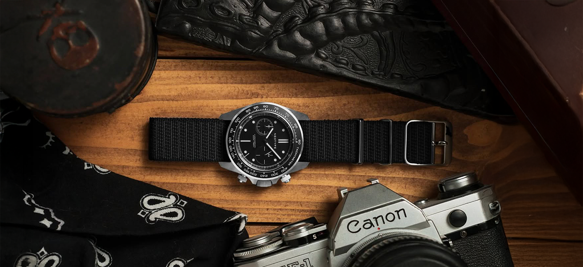 Black single pass watch strap with ladder buckle on chronograph watch laid on wooden surface with camera and accessories.