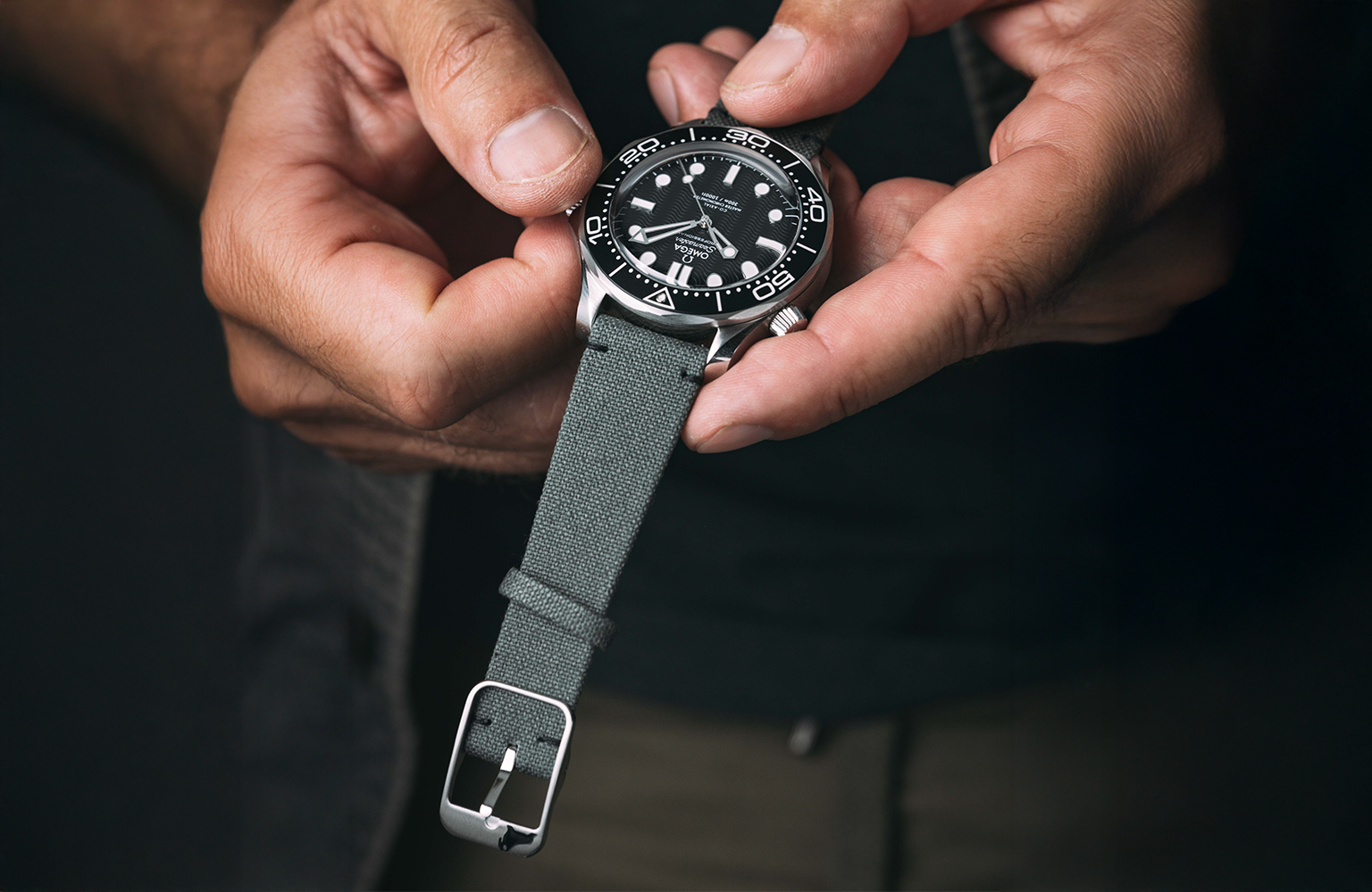 A person holds a wristwatch with a green fabric strap and a black bezel, adjusting the time using the crown. The watch face displays luminous hands and markers.