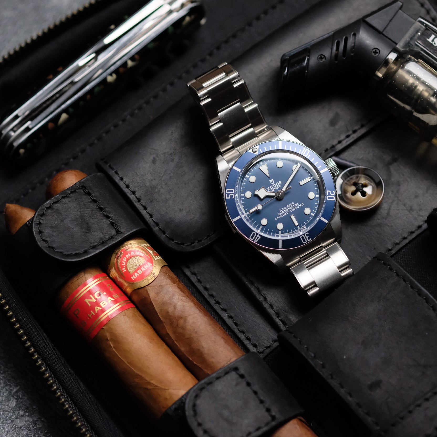 A blue-dial wristwatch with a metal strap is stored in the RSM Watch Strap 3-Piece Travel Watch Case in Black Leather, shown next to two cigars, a pocketknife, and a cigar lighter.