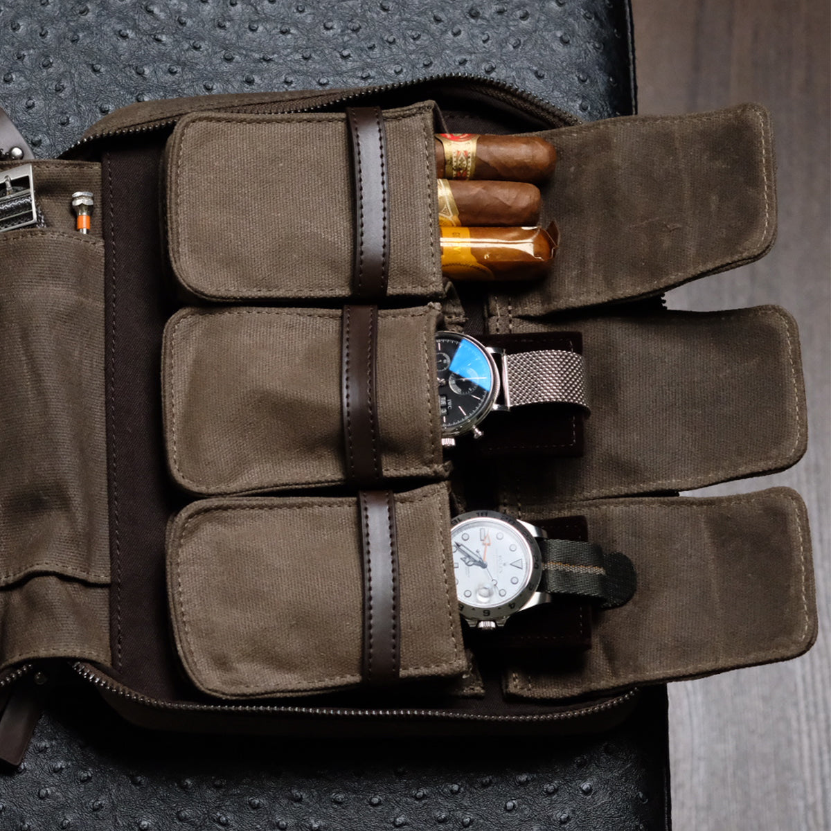 The RSM Watch Strap 3-Piece Travel Watch Case in Waxed Canvas, shown open on a dark textured surface, holds three cigars in one pocket and two wristwatches with different bands and faces in separate compartments.