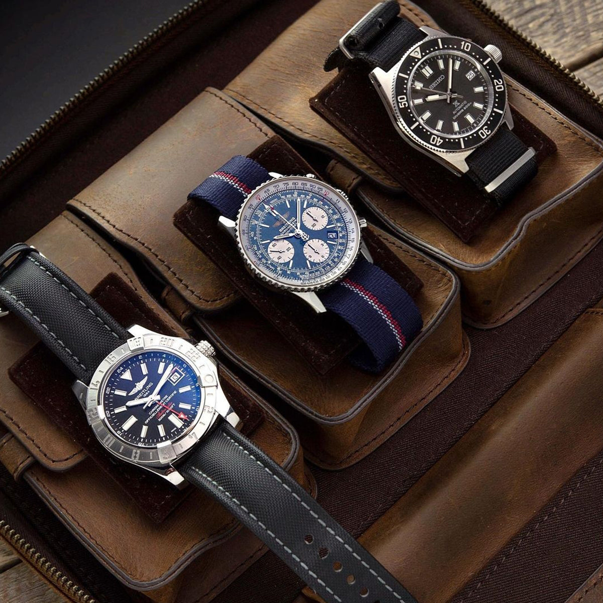 Three watches with black or blue dials rest securely in the RSM Watch Strap 3-Piece Travel Watch Case in Brown Leather—an elegant storage solution displayed on a wooden surface.