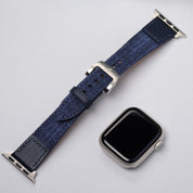 A silver smartwatch with a blank screen is displayed next to the RSM Watch Strap’s Art Deco Silk Watch Strap in Blue, featuring a leaf motif, on a light gray background.
