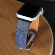 A close-up of a smartwatch with a silver case and an RSM Watch Strap Art Deco Silk Watch Strap in Blue, resting on a round wooden surface.