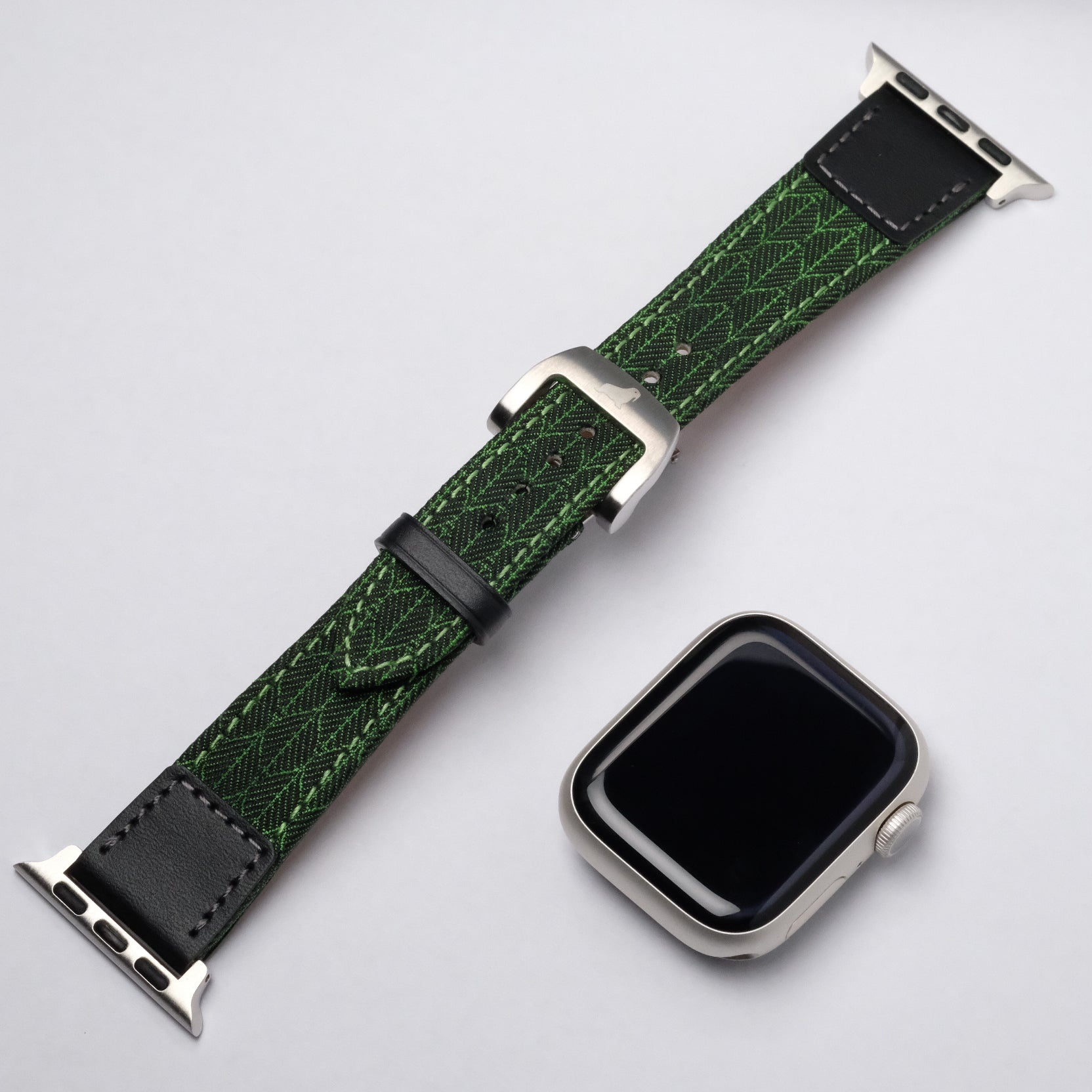 A silver smart watch with a detached RSM Watch Strap—Art Deco Silk Watch Strap in Green, showcasing a geometric leaf pattern, placed on a light gray surface.