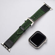 A silver smart watch with a detached RSM Watch Strap—Art Deco Silk Watch Strap in Green, showcasing a geometric leaf pattern, placed on a light gray surface.