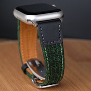 A close-up of a smartwatch with a silver case and black screen, paired with the RSM Watch Straps Art Deco Silk Watch Strap in Green, featuring an Art Deco leaf motif, shown resting on a wooden surface.