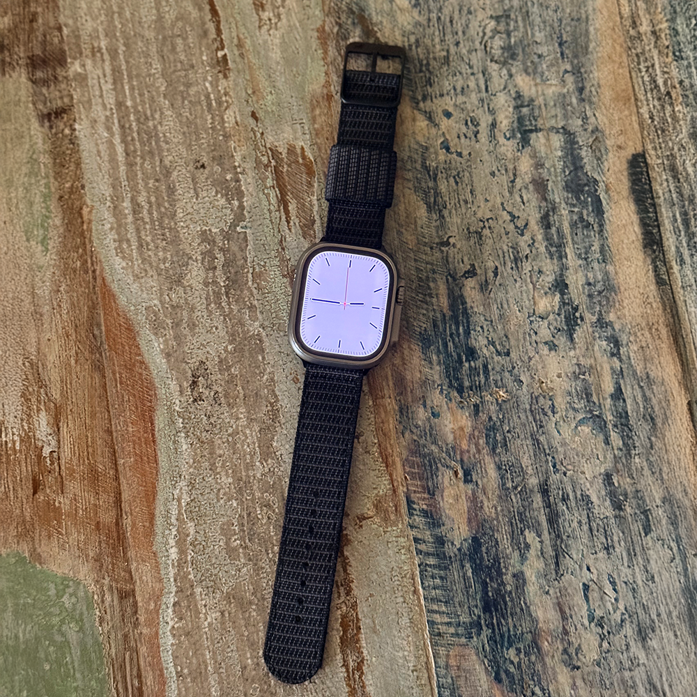 A smartwatch featuring the RSM Watch Strap’s X-Hatch 2-Piece Watch Strap in Black rests on a weathered, multicolored wooden surface. Its display shows an analog clock face with pinkish hour and minute hands.