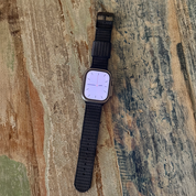 A smartwatch featuring the RSM Watch Strap’s X-Hatch 2-Piece Watch Strap in Black rests on a weathered, multicolored wooden surface. Its display shows an analog clock face with pinkish hour and minute hands.
