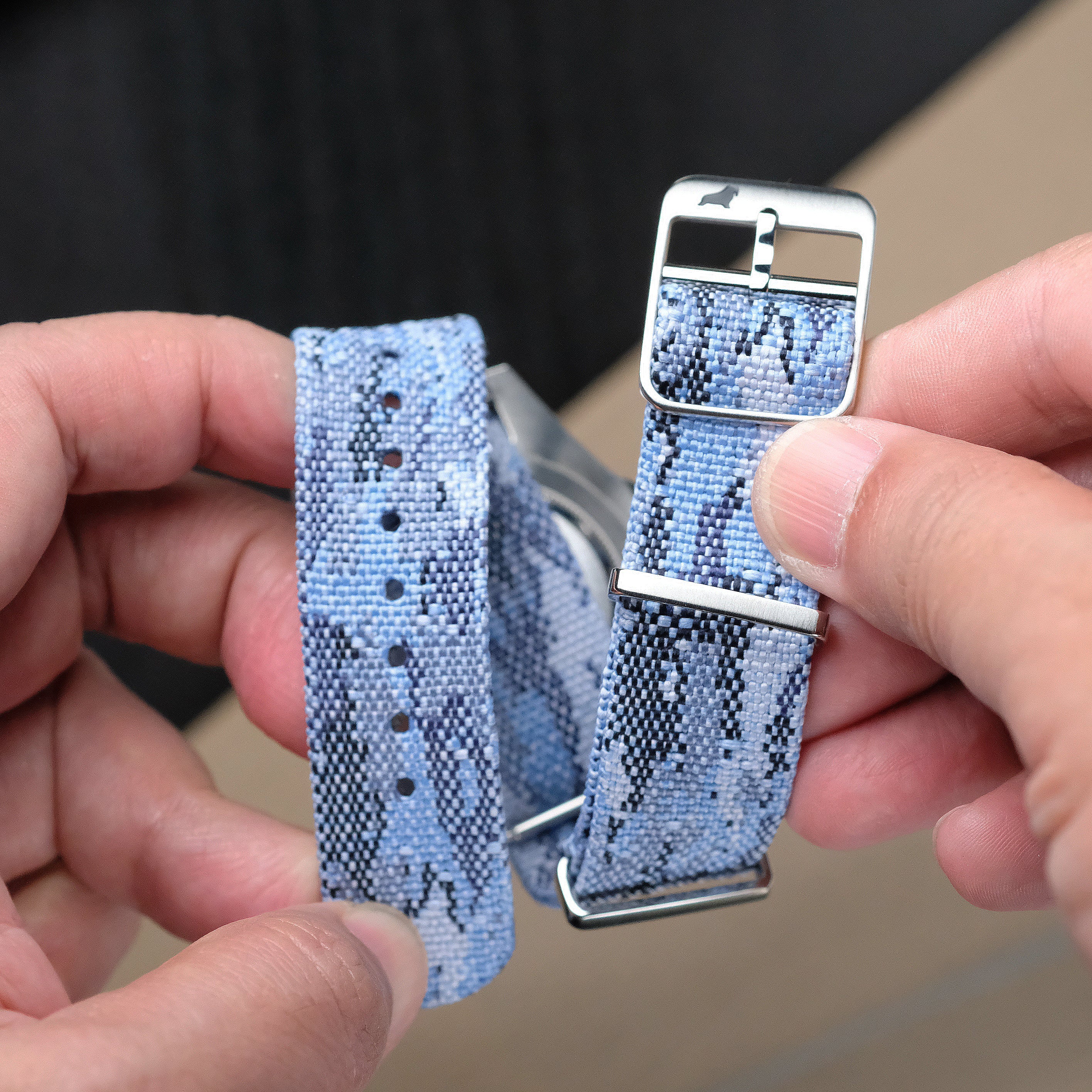 A person holds the RSM Watch Strap Ocean Camo Adjustable Single Pass Watch Strap, featuring jacquard-woven polyester, stainless steel hardware, a silver buckle, textured detail, and adjustment holes.