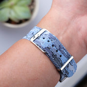 A close-up of a wrist wearing the RSM Watch Strap Ocean Camo Adjustable Single Pass Watch Strap with stainless steel hardware and metal buckle. A blurred green succulent plant is in the background.