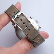 A hand holds an RSM Watch Strap in Taupe, made from Buttero leather with beige stitching and a silver buckle. The watch face appears partially in the background.