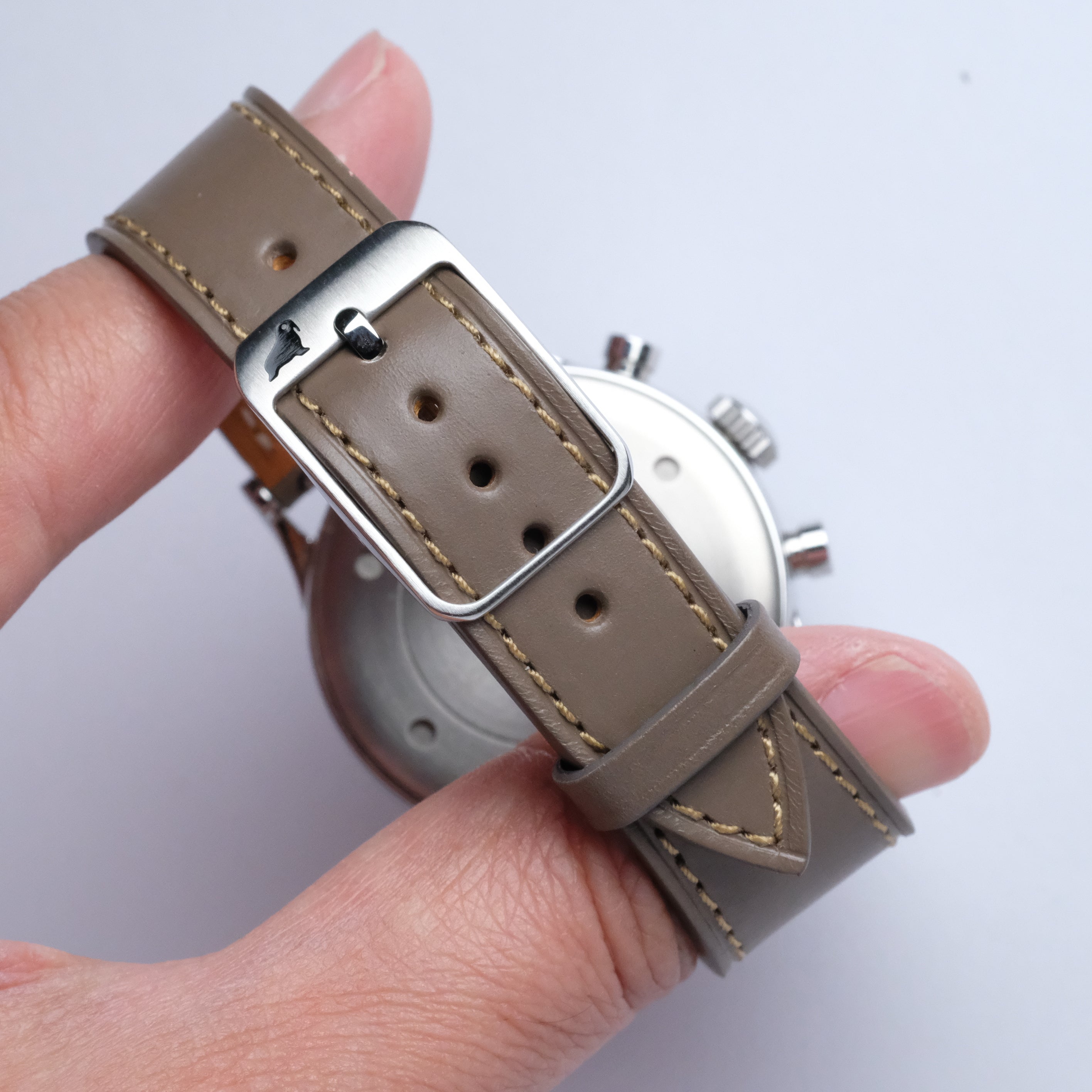 A hand holds an RSM Watch Strap in Taupe, made from Buttero leather with beige stitching and a silver buckle. The watch face appears partially in the background.
