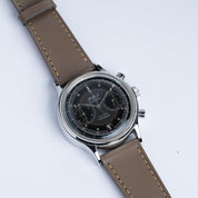 A close-up of a wristwatch with a dark round face, two subdials, silver casing, and an RSM Watch Strap Buttero Leather Watch Strap in Taupe, set against a plain white background.