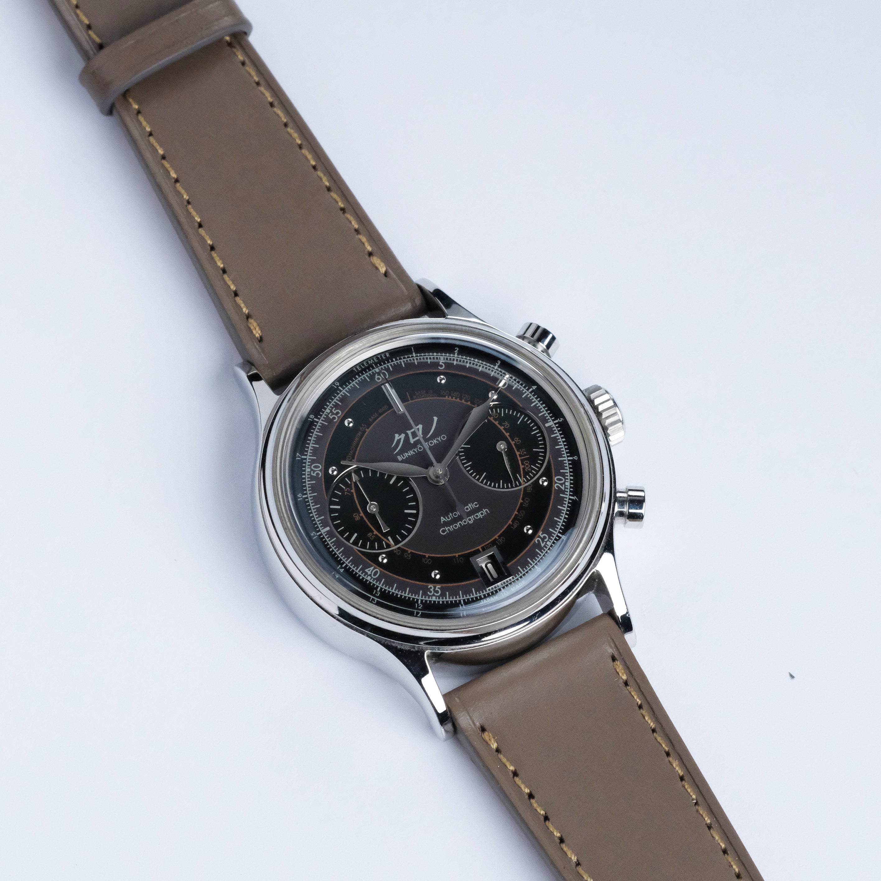 A close-up of a wristwatch with a dark round face, two subdials, silver casing, and an RSM Watch Strap Buttero Leather Watch Strap in Taupe, set against a plain white background.
