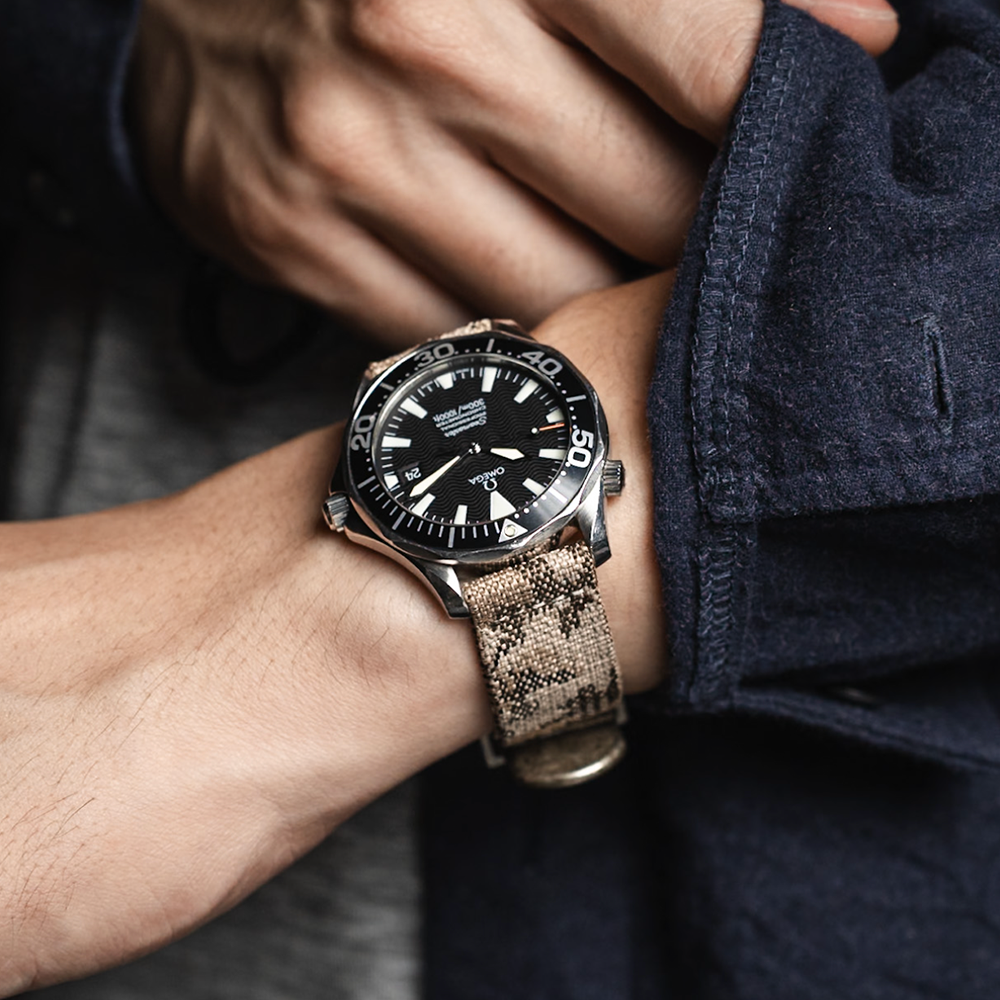 A person adjusts their dark blue jacket sleeve while wearing a wristwatch with an RSM Watch Strap Desert Camo 2-Piece Watch Strap and a black dial.