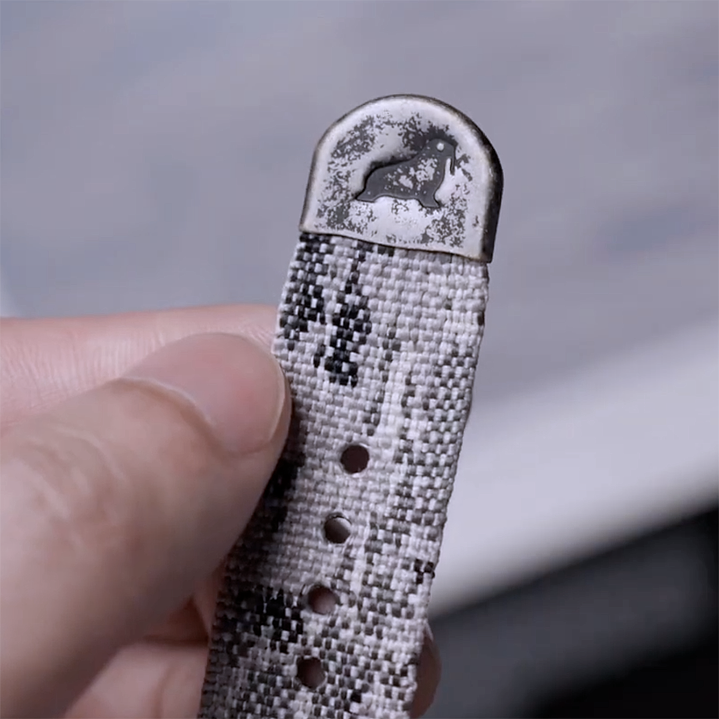 A hand holds the RSM Watch Strap Snow Camo 2-Piece, a jacquard woven watch strap with subtle monochrome camouflage patterns and a metal tip engraved with a prancing horse logo.