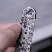 A hand holds the RSM Watch Strap Snow Camo 2-Piece, a jacquard woven watch strap with subtle monochrome camouflage patterns and a metal tip engraved with a prancing horse logo.