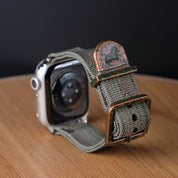 A close-up of the RSM Watch Strap’s Camo Watch Strap, an Apple Watch-compatible gray fabric band with a weathered copper buckle and loop, rests on wood with a black background.