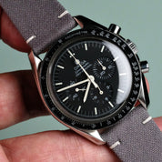A close-up of a hand holding an Omega Speedmaster Professional with a black dial, three subdials, and a Fine Canvas Watch Strap in Mid Grey by RSM Watch Strap. The tachymeter scale is visible on the black bezel.