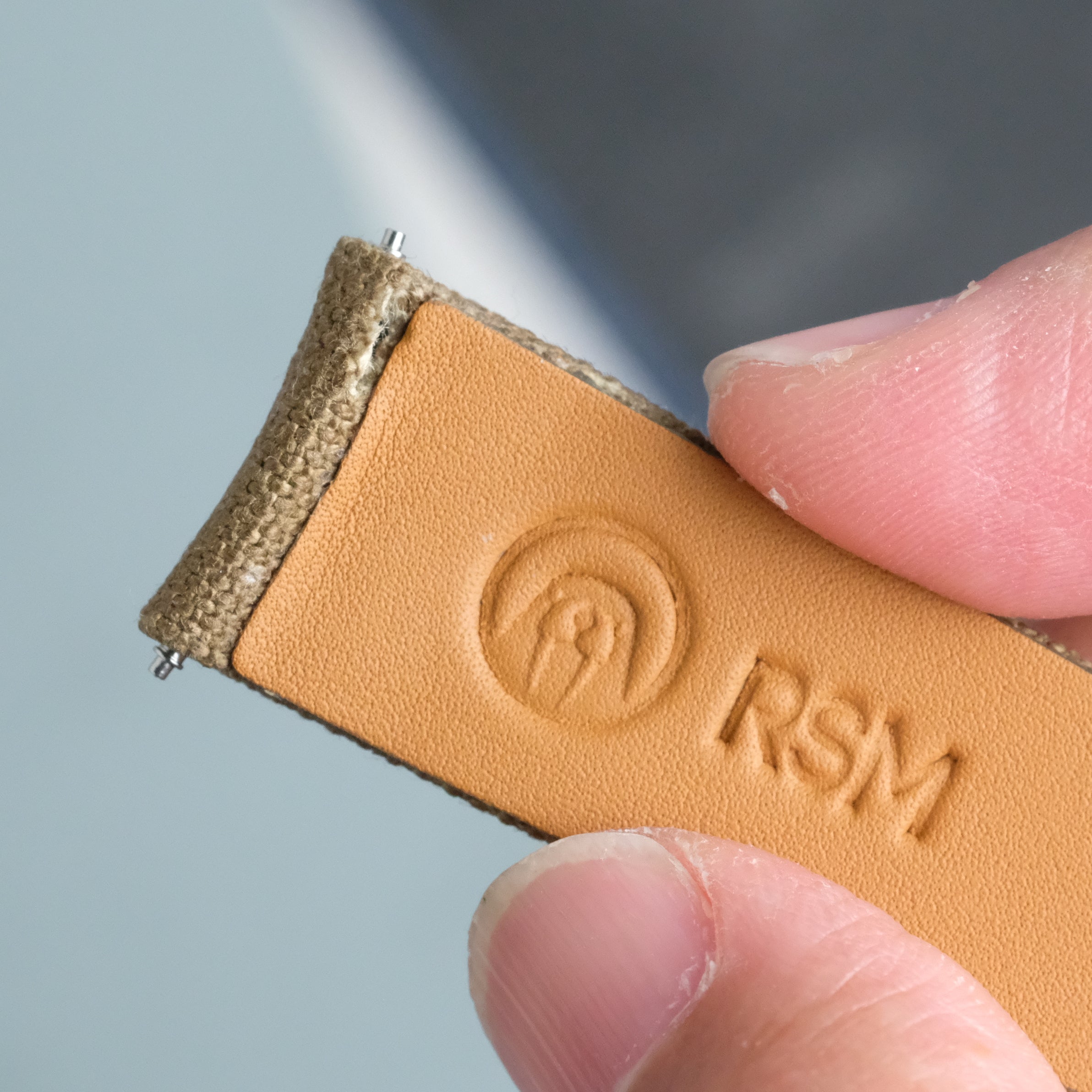 A close-up shows a hand holding an RSM Watch Strap in tan leather with an embossed logo and circular design. Featuring curved spring bars, this strap is ideal for tight lug spaces thanks to its precise stitching and texture.