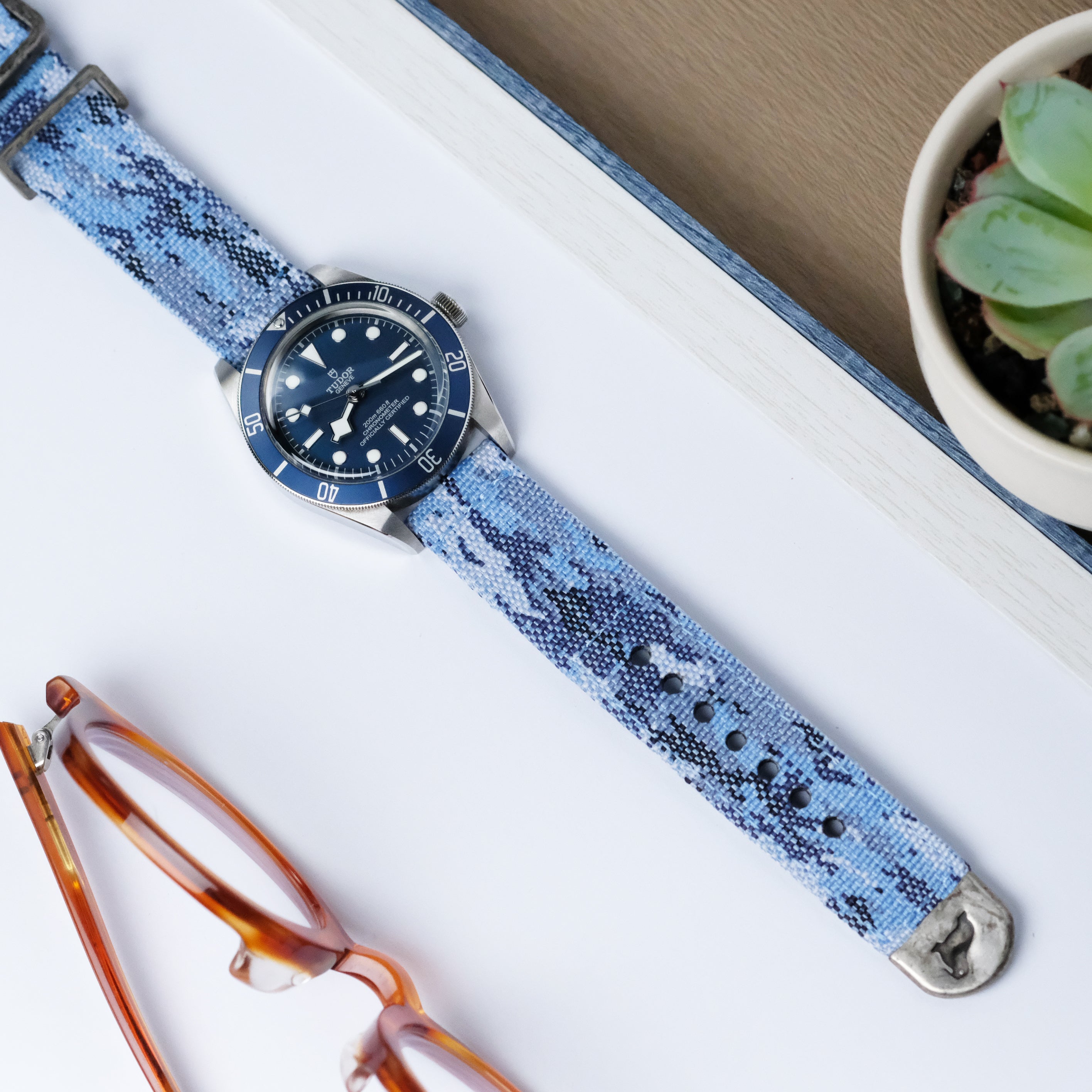 A wristwatch with a blue dial and an RSM Watch Strap Ocean Camo 2-Piece strap sits on a white surface next to brown eyeglasses and a potted succulent.
