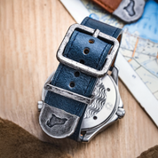 A close-up of the RSM Watch Strap Distressed Leather Watch Strap in Brown features a silver buckle and tip, embossed with a bull, shown beside a map with a watch back displaying a wavy pattern. Limited edition.