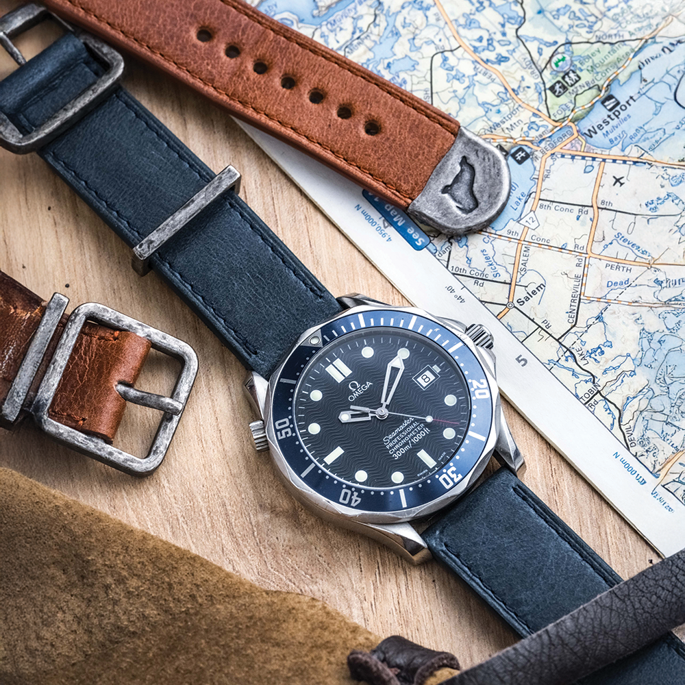 A black Omega wristwatch with a distressed blue leather strap rests on wood beside an RSM Watch Strap Distressed Leather Watch Strap in Brown and a map. The watch face displays 10:10.