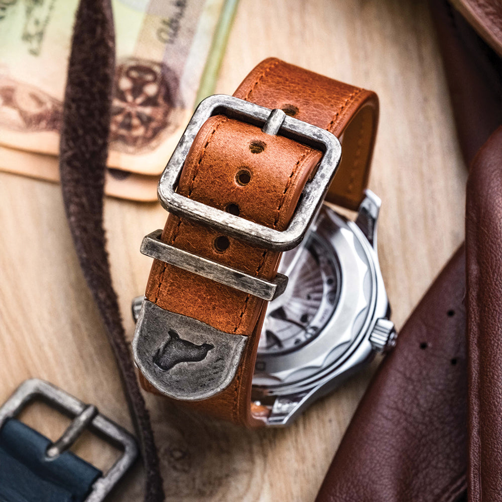 The Distressed Leather Watch Strap in Brown by RSM Watch Strap, featuring a rustic metal buckle and bull head engraving, rests on a wooden surface near a watch, leather accessories, and partially visible currency notes.