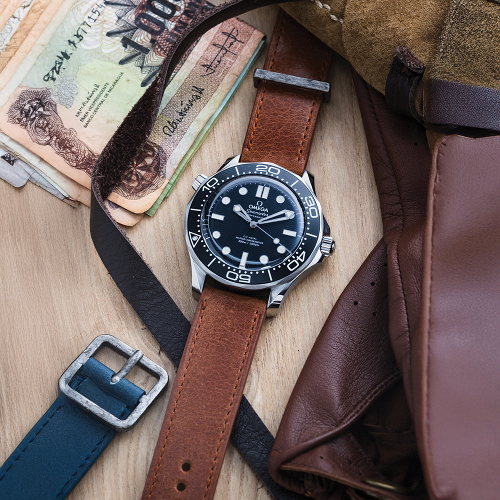 A RSM Watch Strap Distressed Leather Watch Strap in Brown is displayed on a wooden surface alongside leather gloves, paper currency, and a blue belt buckle.
