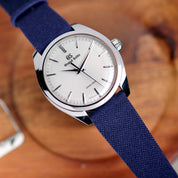 A Grand Seiko wristwatch with a silver sunburst dial, silver hour markers, and an RSM Watch Strap Fine Canvas Watch Strap in Navy Blue is set on a wooden surface. The watch features a polished stainless steel case.