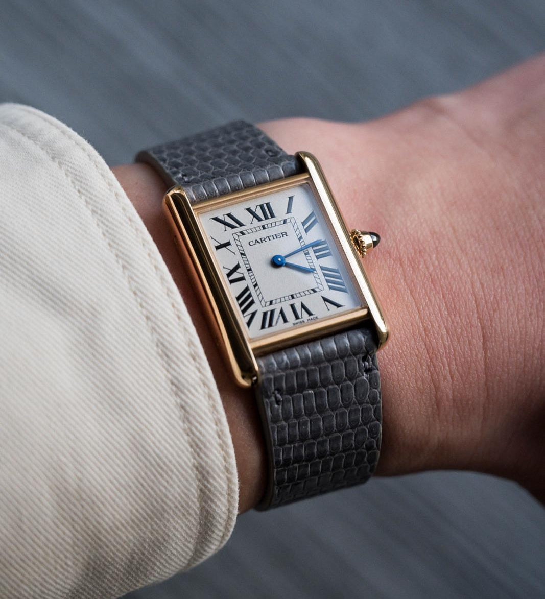 A close-up of a wrist wearing a Cartier Tank watch with a gold rectangular case, white dial, black Roman numerals, and blue hands, paired with an RSM Watch Strap Lizard Leather Watch Strap Vertical Grain in Grey. Light-colored sleeve visible.
