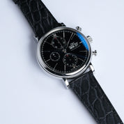 The RSM Watch Strap Alligator Circular Grain Calf Leather Watch Strap in Black is shown on a luxury wristwatch with a black dial, three subdials, date and day display, and silver case against a white background.