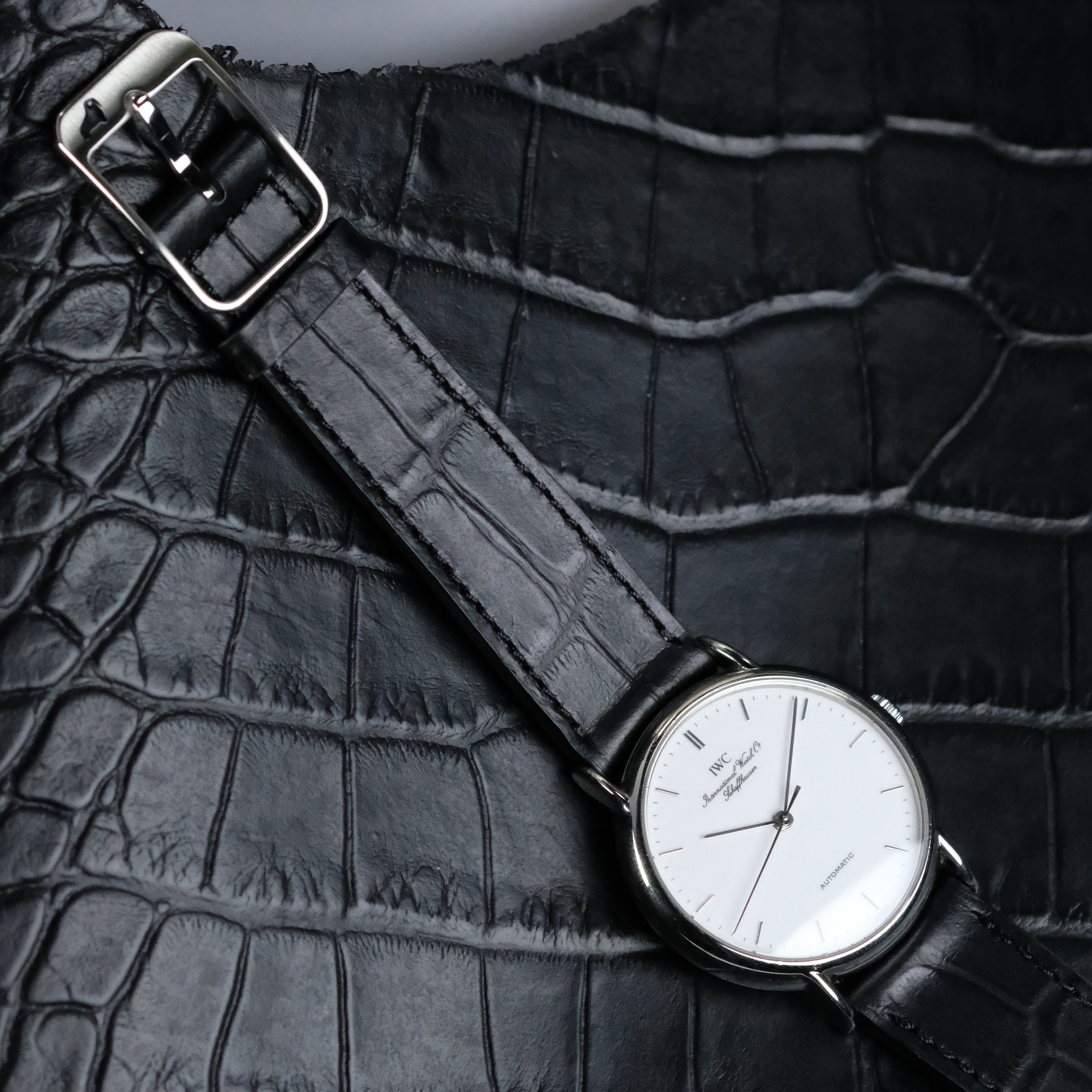 The RSM Watch Strap Curved Lugs Alligator Rectangular Grain Watch Strap in Black, paired with a white-dial wristwatch, rests on textured black leather, highlighting refined Japanese craftsmanship.