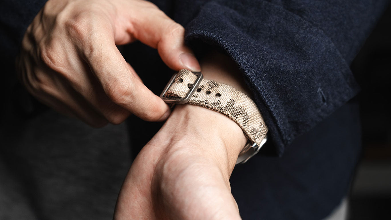 Close-up of an RSM desert camouflage watch strap being adjusted on the wrist, showing woven texture, camo pattern, and brushed metal buckle.