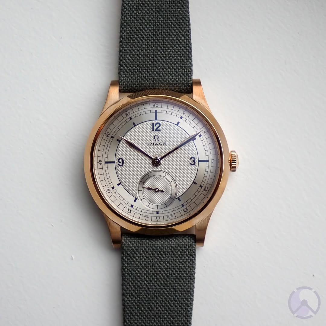 A gold Omega wristwatch with a silver dial, blue hands, black numerals, and textured center features a small seconds subdial at 6 o’clock and is paired with the RSM Watch Strap Fine Canvas Watch Strap in Brunswick Green, shown on a white background.