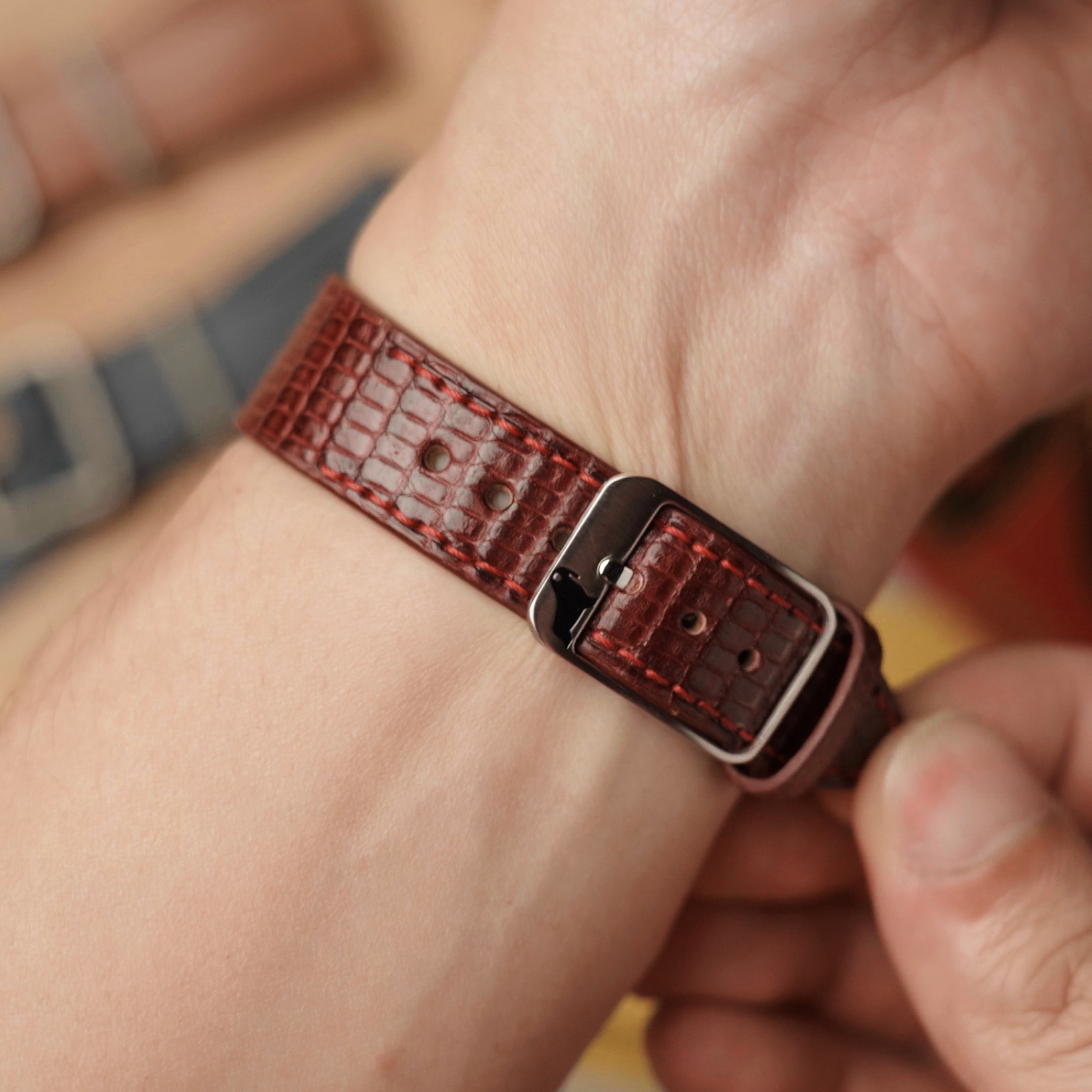 Person adjusting RSM's red lizard leather watch strap on a wrist with a blurred background