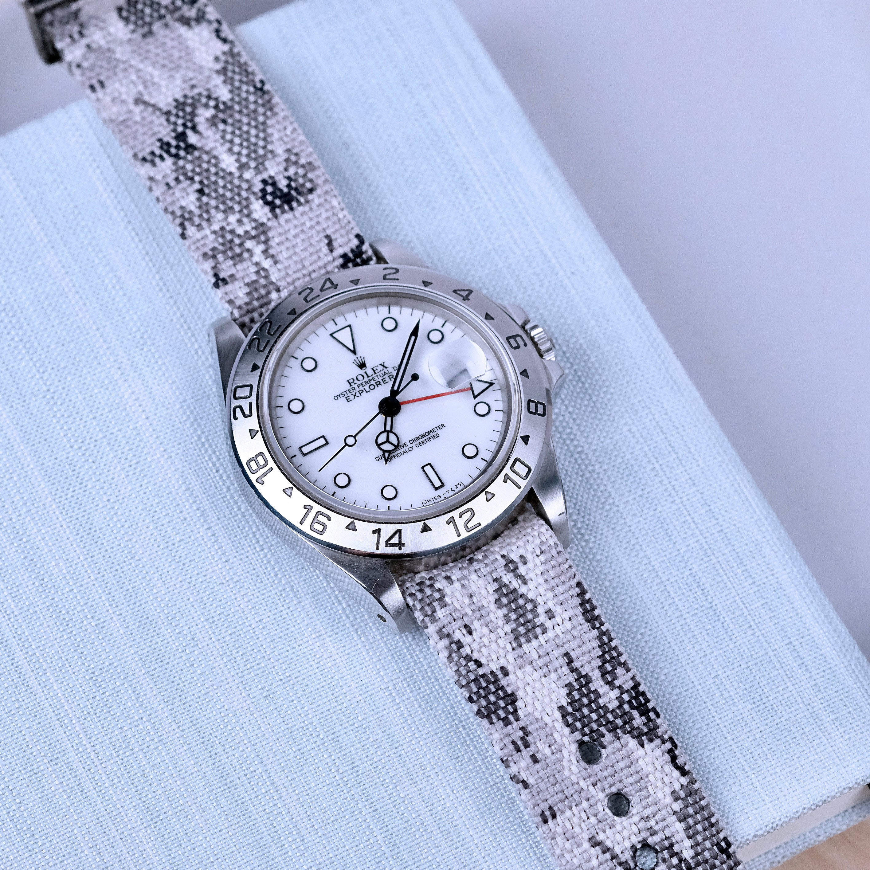 A silver wristwatch with a white face and black hour markers rests on a light blue textured surface, featuring a bezel with 24-hour markers and an RSM Watch Strap Snow Camo Zulu watch strap in monochrome camouflage for added style.
