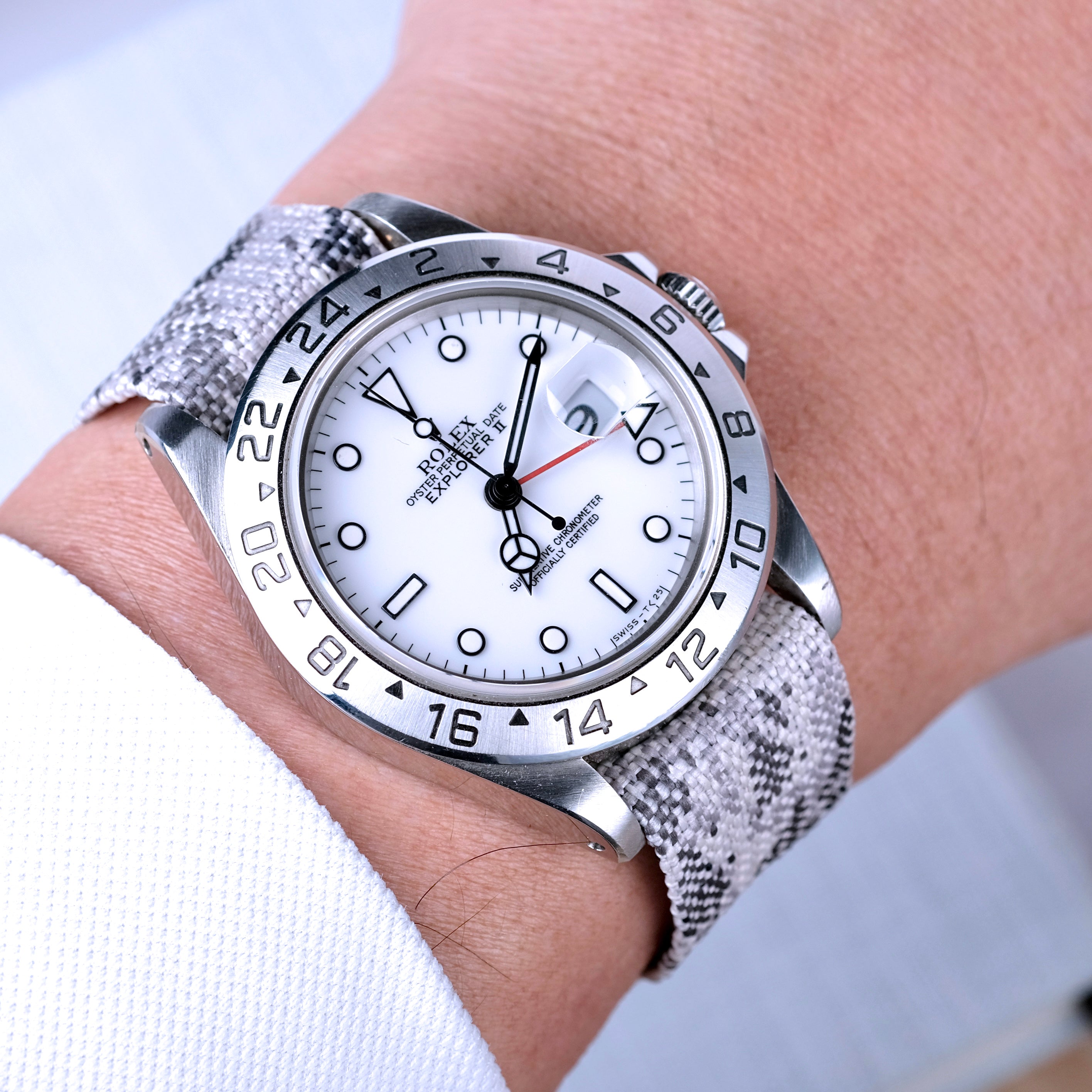 A person wearing a white long-sleeve shirt sports a silver wristwatch with a white dial, black markers, date window, 24-hour bezel, and an RSM Watch Strap Snow Camo Adjustable Single Pass jacquard woven strap.