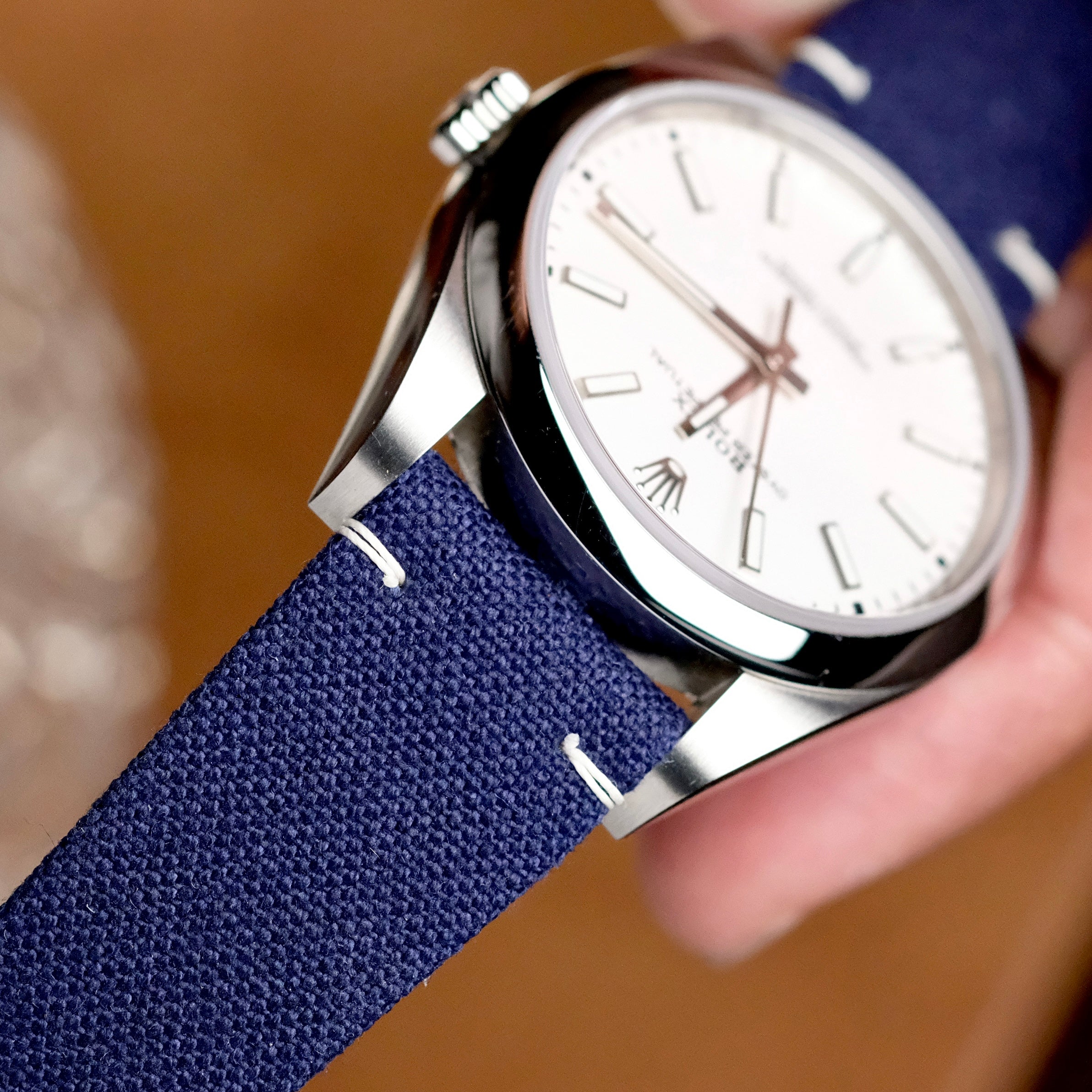 A close-up of a wristwatch featuring a white dial, silver case, and the RSM Watch Strap’s Fine Canvas Watch Strap in Navy Blue. The watch is held at an angle by a hand, showcasing its hour markers and stylish design.